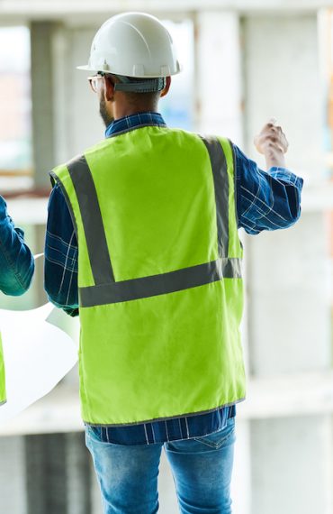 Back view portrait of two construction workers wearing hardhats and reflective vests discussing engineering plans on site, copy space